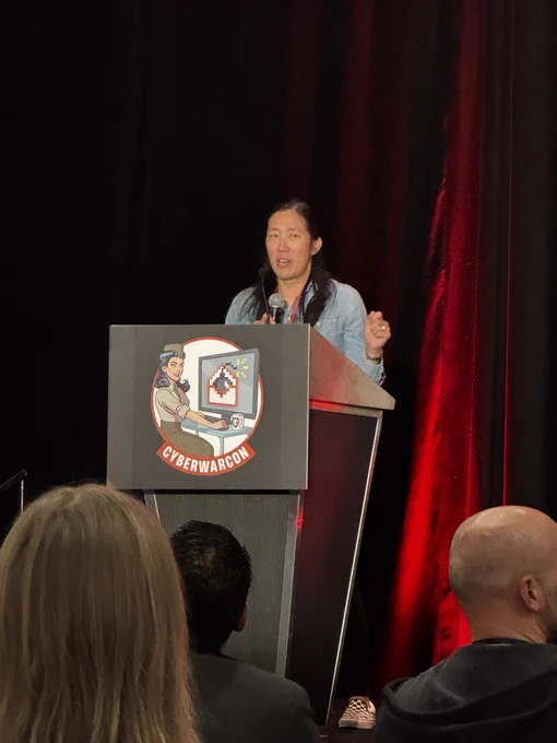 Judy Ng stands on stage behind a podium, with the CYBERWARCON logo on it.