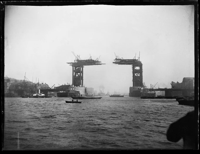 A black and white photograph of a bridge under construction over a river.