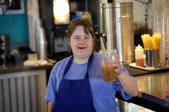 Kristen in the cafe that she works at holding up an iced coffee and smiling.