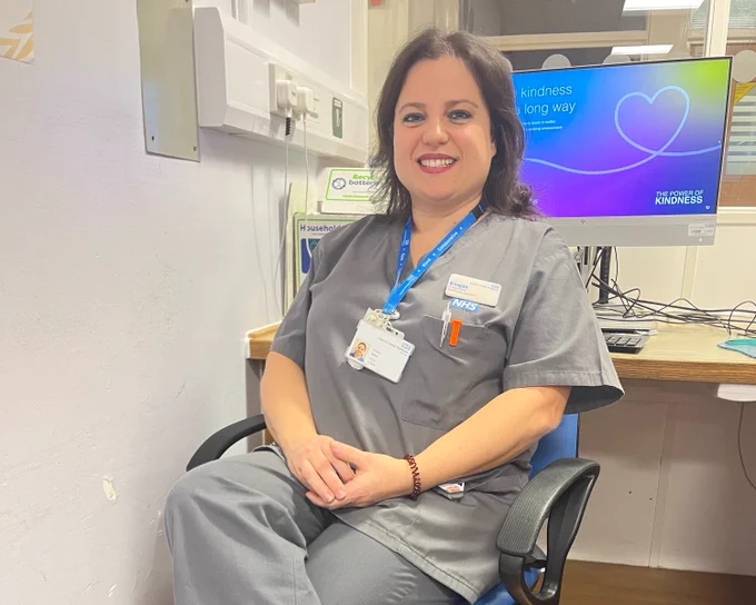Close up photo of a support worker at Imperial College Healthcare. They are wearing a grey NHS uniform, sitting at a desk and smiling towards the camera.