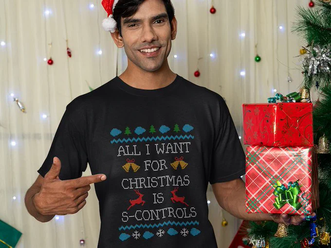 A man holding some wrapped gifts, standing next to a Christmas tree. He is wearing a t-shirt with a faux knit design on. The design has images of blue clouds, Christmas trees, bells, and snow-flakes. The t-shirt also has the words "All I want for Christmas is S-Controls" .