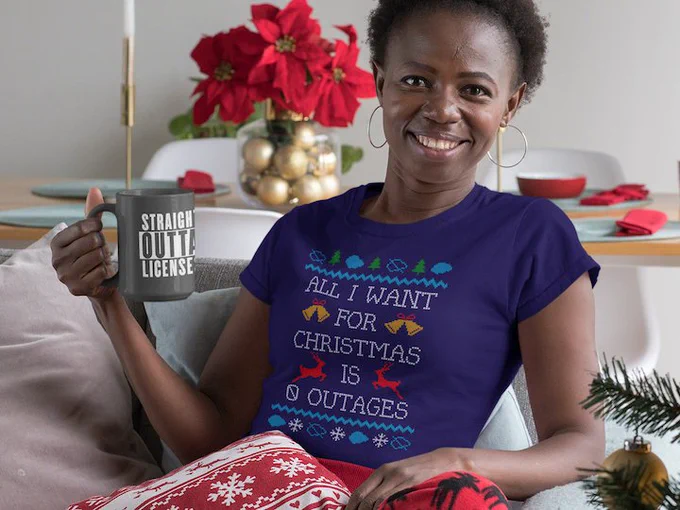 Lady sitting on a sofa with a mug of hot drink. There are Christmas decorations in view, and she's wearing a festive faux-knit t-shirt with images of blue clouds, Christmas trees, bells, and snow-flakes. The t-shirt also has the words "All I want for Christmas is 0 outages on it"