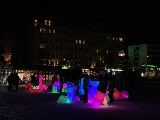 A wide angle view of Twist and Shine in Hämeenlinna Market Square in the city centre. There are about 20 sculptures of different sizes, and dozens of people playing with them, surrounded by buildings.