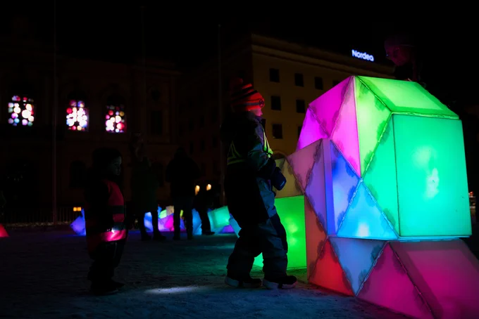A child approaches a structure composed of two large sculptures stacked on top of each other, creating a colourful, geometric wall. The lights are reflected in the white snow underneath them, and more glowing sculptures are visible in the background.