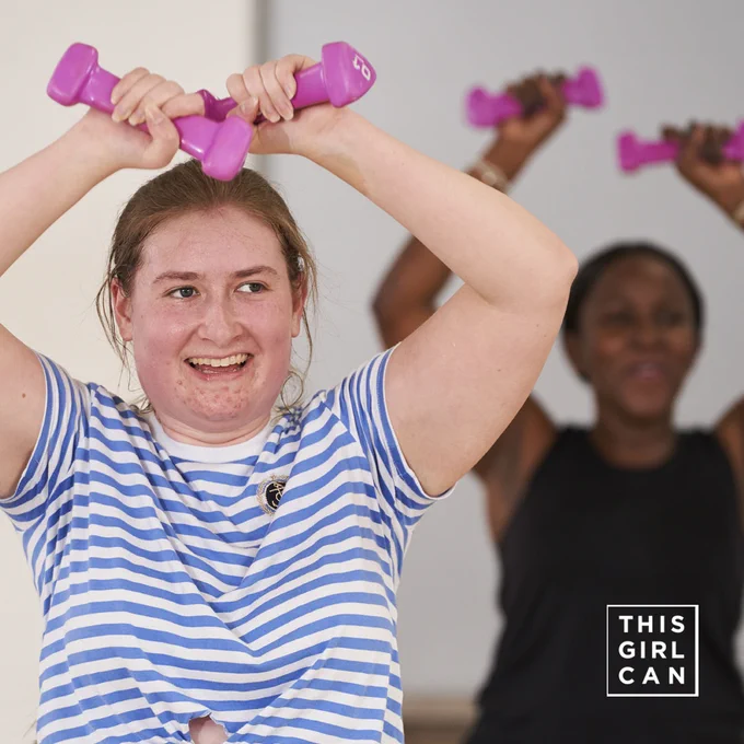 Person in a striped shirt lifting dumbbells with a smiling expression and another person visible in the background. There is a "This Girl Can" campaign logo.