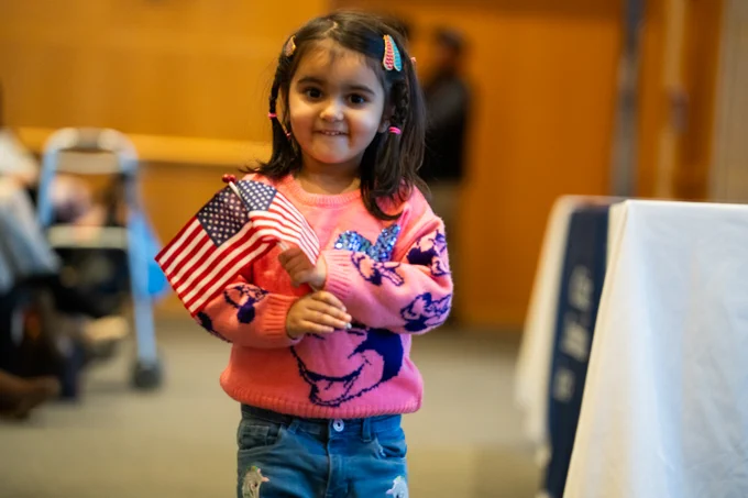 A child holding two American flags smiles in a room, wearing a pink sweater with a heart design and jeans.