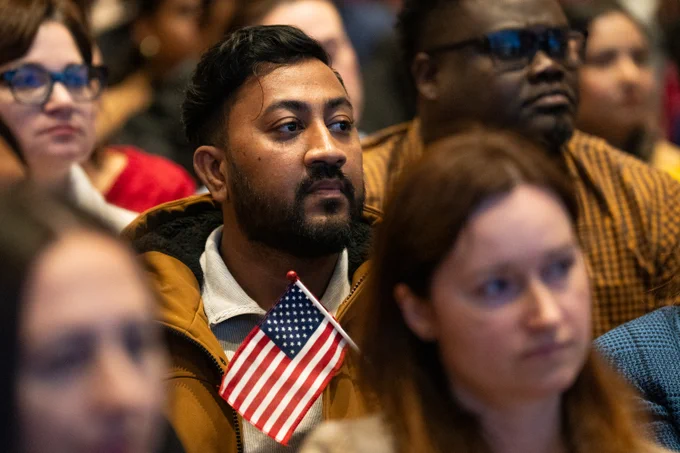 A diverse audience attentively listening at an naturalization ceremony at the JFK Library, with one person holding a small American flag.