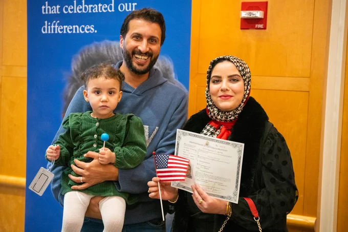 Family at a U.S. citizenship ceremony at the JFK Library. An adult is holding a toddler while another adult displays a naturalization certificate and a small American flag.