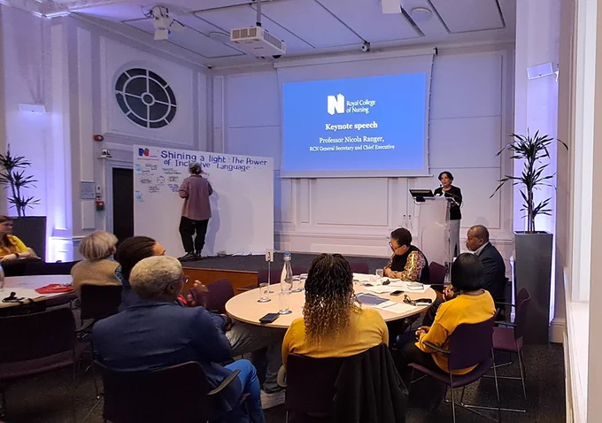 A presentation at the Royal College of Nursing featuring a keynote speech by its General Secretary and Chief Executive, Professor Nicola Ranger. Multiple attendees are seated around tables, listening attentively in a room decorated with potted plants and promotional banners.