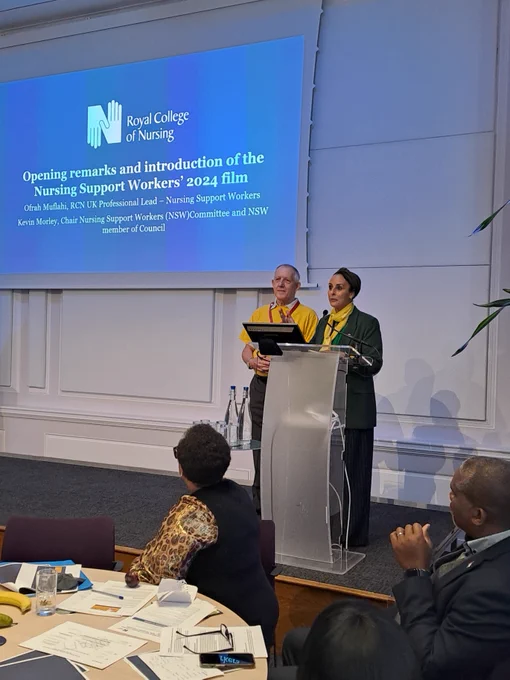 Ofrah Muflahi, RCN Professional Lead for Nursing Support Workers, speaking at a podium during the Royal College of Nursing Support Workers' Conference. Behind her is a digital, screen titled "Opening remarks and introduction of Nursing Support Workers' 2024 film" Audience members are seated, listening attentively.