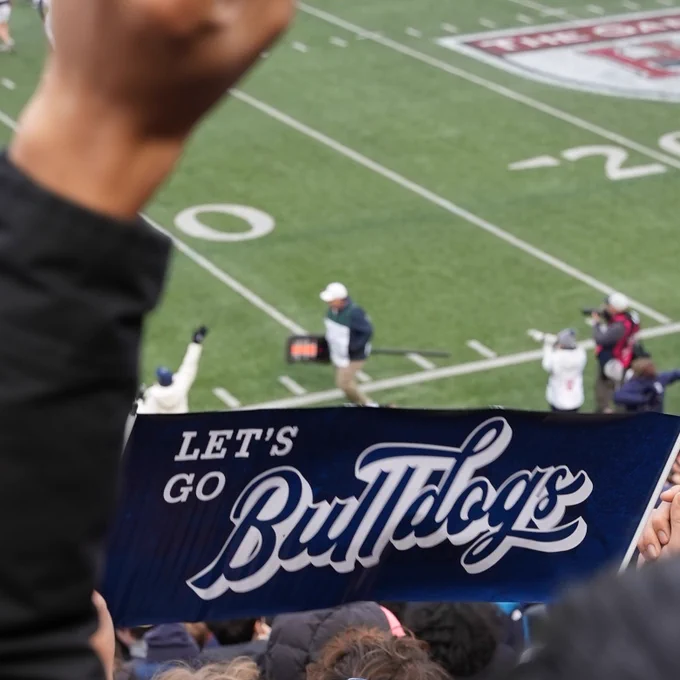 A person holding a "Let's Go Bulldogs" sign at a crowded sports event, with a coach and players in the background on a football field.
