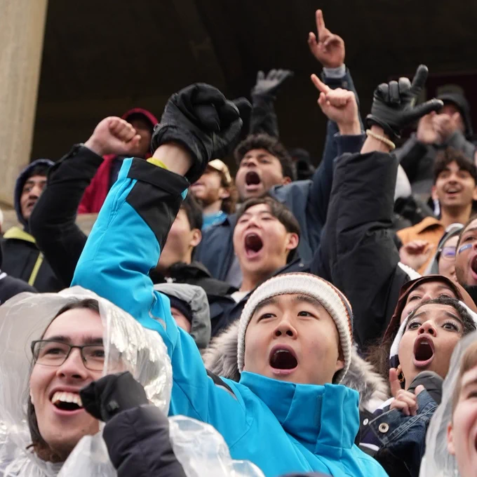 Excited crowd of people cheering at a sports event, some wearing rain ponchos, with one person in a bright blue poncho pointing and shouting enthusiastically.
