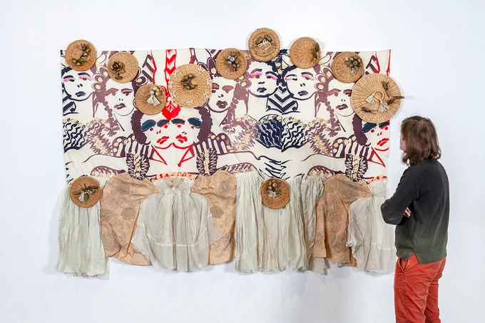 A visitor observes a large artwork composed of painted dancers, fabric, and basket-lid like objects arranged rhythmically.