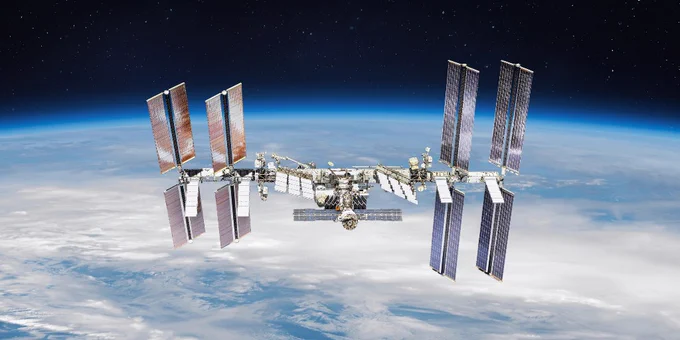 The International Space Station orbits Earth, with large solar panels extended on either side, set against the backdrop of space and Earth's atmosphere.
