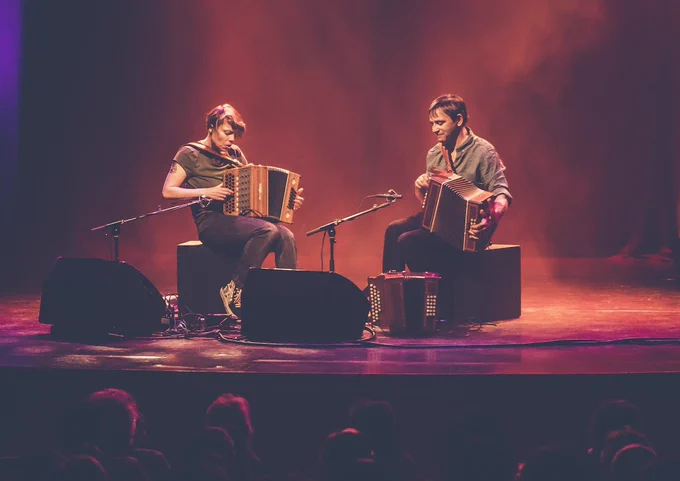 Photo of Andy Cutting and Anne Niepold sat beside each other on stage. They're both focused on playing their diatonic accordions, and an audience is watching on, no doubt spellbound!