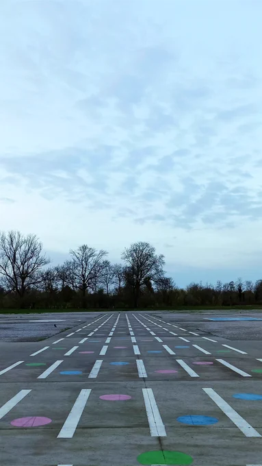 Sous un ciel gris bleu parsemé de nuages, grande esplanade de béton sur laquelle sont dessinées sept lignes discontinues parallèles. Entre ces lignes, de gros points ont été représentés. Ils sont alternativement roses et bleus. Au premier plan le rond est vert et est dans l'axe de la ligne centrale. En arrière plan, la limite du tiers inférieur de la photo est occupée par une végétation dense constituée d'arbres plus ou moins hauts. 