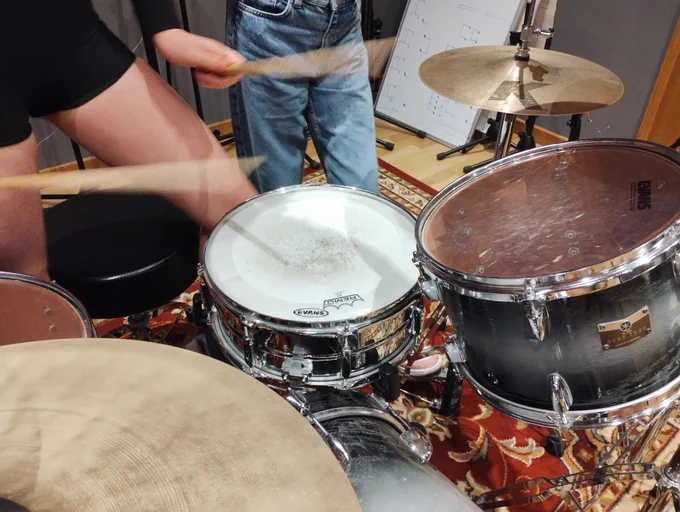 Young person playing drums with other person in background watching