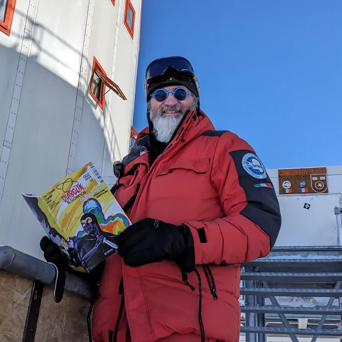 Un uomo vestito con giacca per temperature artiche e occhiali da sole guarda in camera. In mano, tiene un fumetto dalla copertina gialla: si tratta di The Climate Change Issue, il crossover tra Diabolik e Comics&Science. La persona è Alberto Salvati, ingegnere informatico del CNR e membro del team presso la base Concordia in Antartide. Alle sue spalle uno scorcio della base e un cielo terso.