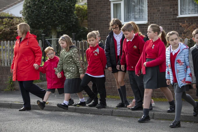 Mae grŵp o blant sy'n gwisgo gwisg ysgol goch yn croesi'r ffordd gydag athro.