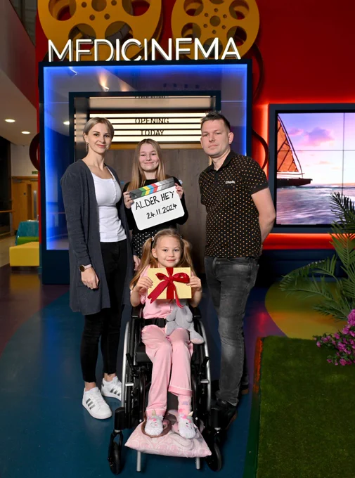 Young girl in a wheel chair and her family standing behind her are smiling into the camera whilst stood in front of the entrance to the MediCinema at Alder Hey Children's Hospital. 