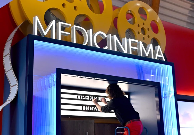 A man is up a ladder arranging letters in a cinema light box which reads 'Opening Today. Above the light box is a neon sign which reads medicinema