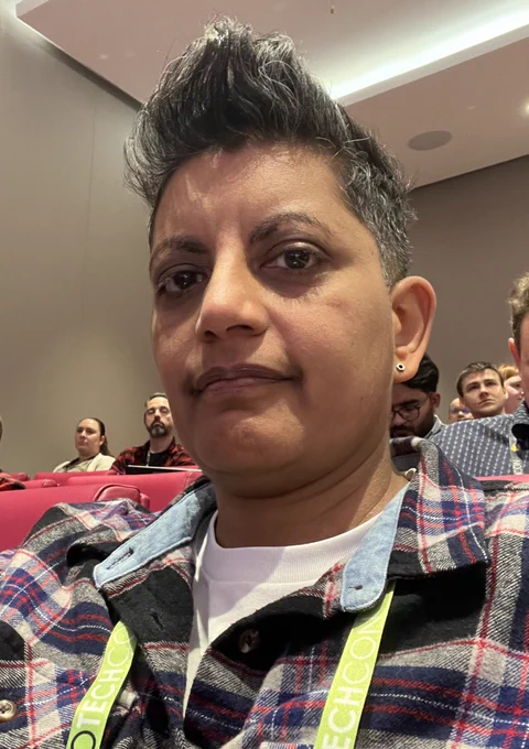 Head shot of me at tech con. Ihave short  salt and pepper hair and brown skin I’m wearing a check shirt in dark blue and red with a white shirt under l. I have a green lanyard on.