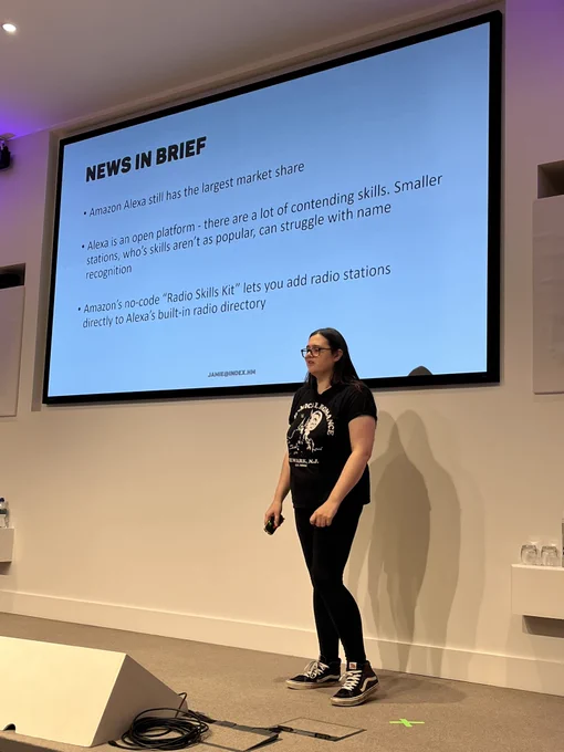 NEWS IN BRIEF
• Amazon Alexa still has the largest market share
• Alexa is an open platform - there are a lot of contending skills. Smaller stations, who's skills aren't as popular, can struggle with name recognition
• Amazon's no-code "Radio Skills Kit" lets you add radio stations directly to Alexa's built-in radio directory

Jamie is on stage in front a slide with the text above. They are dressed in black and has long hair and glasses and van trainers.