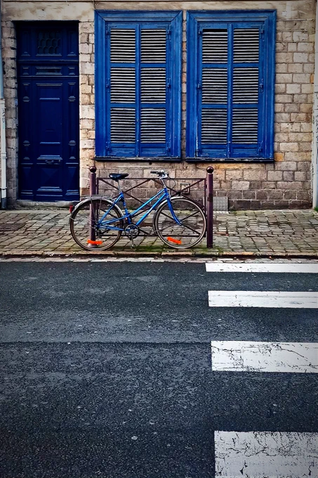 Cette photo montre un décor urbain élégant et pittoresque, centré sur une façade en pierre ornée de volets et d'une porte bleu profond. Les volets sont fermés, et leur peinture écaillée suggère un charme ancien ou une histoire vécue. Un vélo bleu, assorti à la façade, est attaché à une barrière en métal marron, créant une harmonie visuelle entre les éléments. La scène est encadrée par une chaussée et un passage piéton au premier plan. 