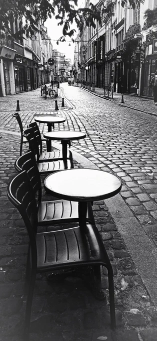 Cette photographie en noir et blanc capture une rue pavée, déserte et baignée d'une lumière douce du matin. Au premier plan, des tables rondes et des chaises alignées créent une ambiance typique de terrasse de café, mais sans aucun client. La perspective de la rue s'étire vers l'horizon, encadrée par des immeubles de style ancien et des vitrines de magasins. Des vélos attachés à un poteau et des enseignes suspendues ajoutent des détails urbains subtils à cette scène calme et mélancolique.