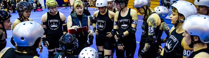 Gotham City Roller Derby team are wearing black vests and silver helmets while a female coach reads something off a clipboard.