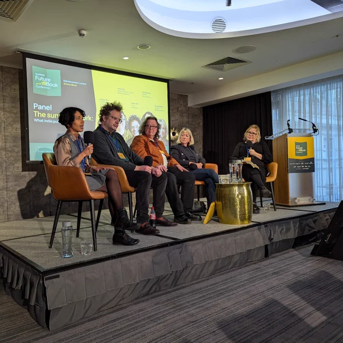 five panelists on stage, sat on chairs holding hand held microphones