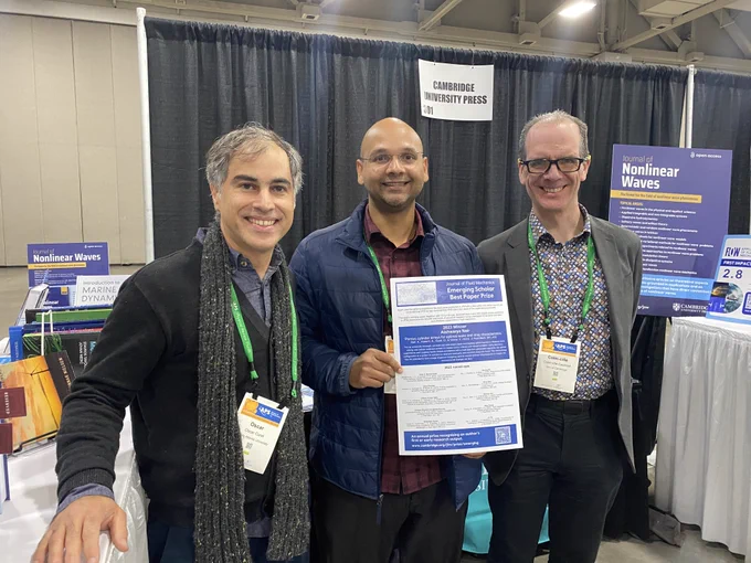 Oscar Curet and Siddhartha Verma standing with Colm-cille Caulfiled at the APS-DFD Cambridge University Press exhibition booth, one holding a certificate promoting teh Emerging Scholar best Paper Prize. They are smiling and positioned in front of promotional banners.