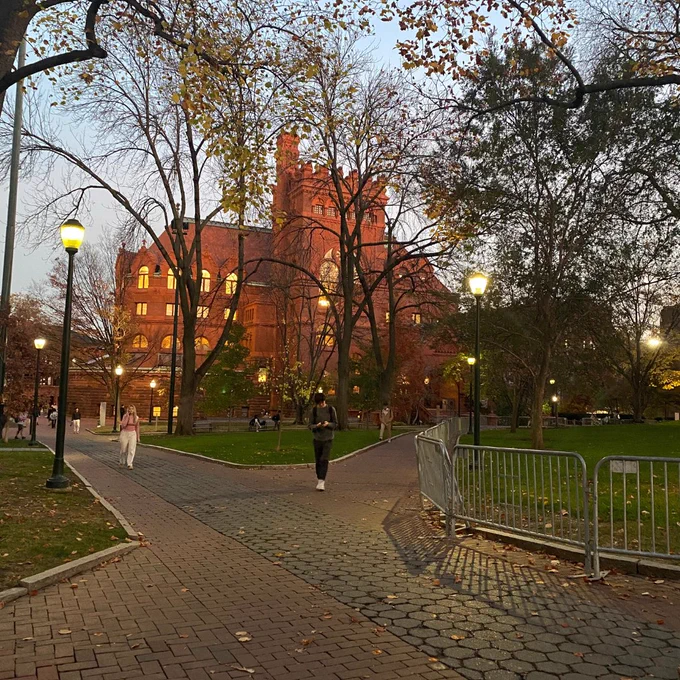 A person walks along a pathway on a university campus at dusk, with the illuminated, historic red brick building in the background, surrounded by autumn foliage and lampposts.
