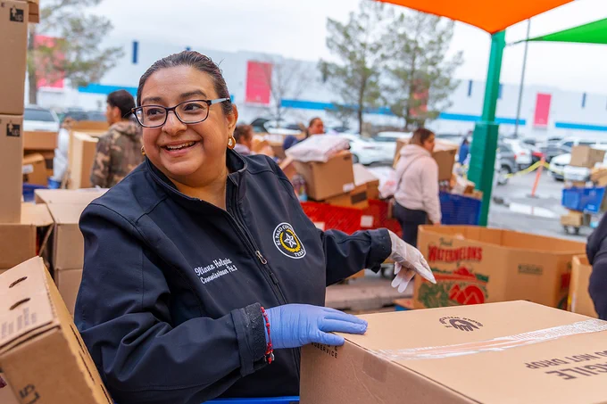 A woman in a navy blue jacket and gloves handling a box at a community service event under a tent.