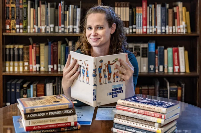 Photo of library staff holding a cookbook with books behind her and on a table around her