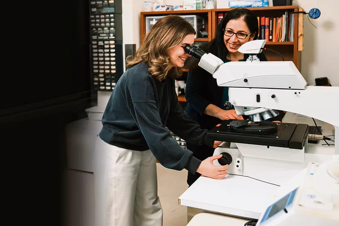 A person looking through a microscope while a second person looks on smiling