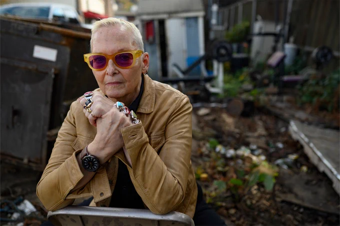 Individual with sunglasses and a leather jacket sitting outdoors with their elbows on their knees, surrounded by an urban backdrop with dumpsters and debris.