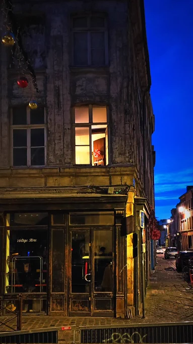 La photo capture une scène matinale, à l'angle de deux rues. Une fenêtre éclairée au premier étage attire l’attention avec une décoration en forme de grosse sucette rouge et blanche, qui rappelle le motif visible sur l’enseigne située dans la rue à droite. En bas, une boutique au style vintage, sombre et rustique, à la devanture en bois patiné, contraste avec la lumière douce émanant de la fenêtre. La rue pavée est bordée de bâtiments anciens éclairés apr des lampadaires, et les boules de Noël suspendues ajoutent une touche festive à l’ensemble. L’éclairage bleu profond du ciel nocturne donne une atmosphère chaleureuse et hivernale à la scène.

