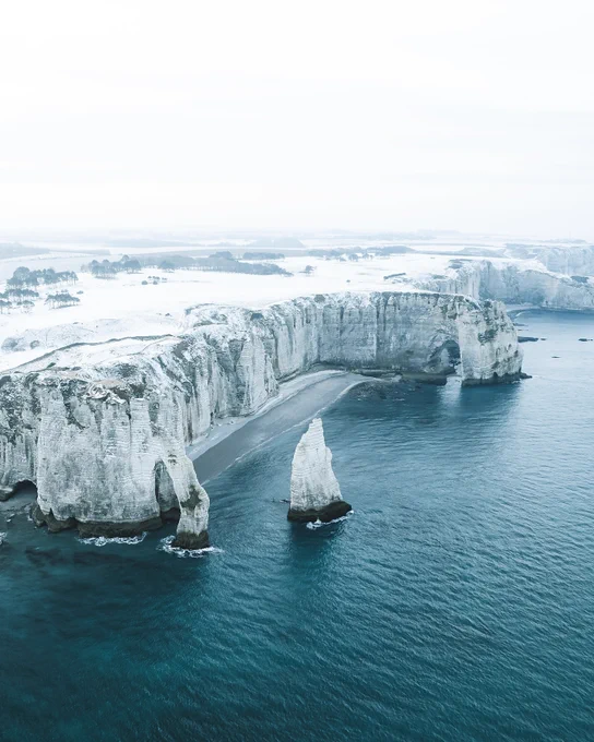 Photo des falaises d'Etretat enneigées. Le tiers du bas de l'image est occupé par la mer, d'un très beau bleu froid, puis viennent les rochers gris qui y plongent, le plateau couvert d'un manteau blanc et de quelques arbres, et enfin le ciel d'un blanc laiteux