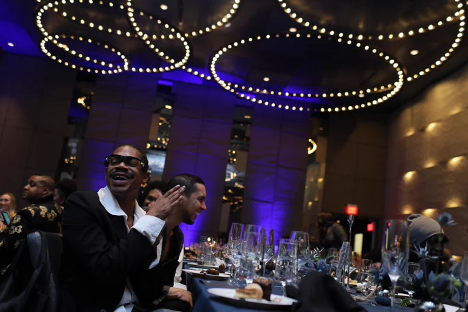 A photo from the 2023 Luminary Gala of Telfar Clemens seated a table. He is smiling and clapping.