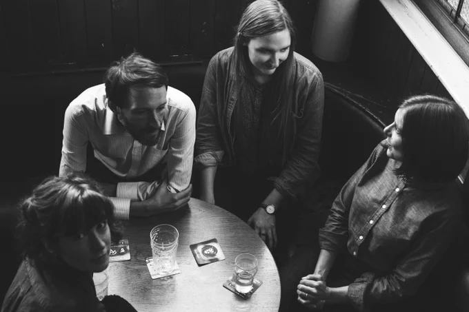 Black and white photo of The Furrow Collective sat around a table at a pub, with a few beer mats and a couple of empty glasses in front of them. Emily and Rachel are chatting, Alasdair looks thoughtful and Lucy is looking up towards us. 
