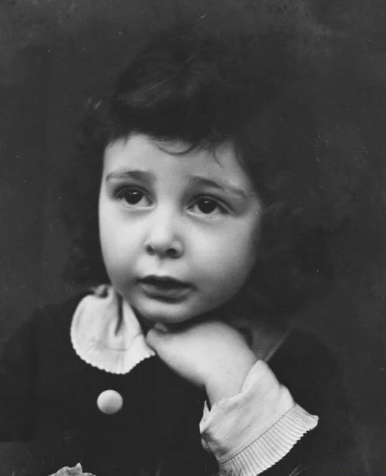 A young boy with long dark hair photographed in a dark blouse with large white collar. His left hand is under his chin. 