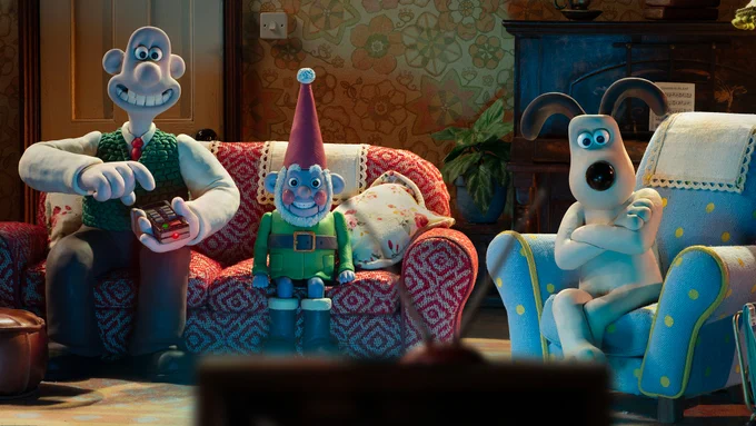 Wallace and Gromit watch television with Norbot, a smart gnome Wallace has created. Wallace holds the television remote and sits on the sofa with Norbot while Gromit sits in an armchair with his arms folded. They're all looking at a television.