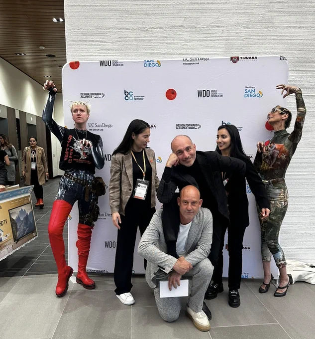 The team poses in front of the conference's sponsor backdrop. Some are kneeling, raising their arms dramatically, smiling, and/or leaning on each other.