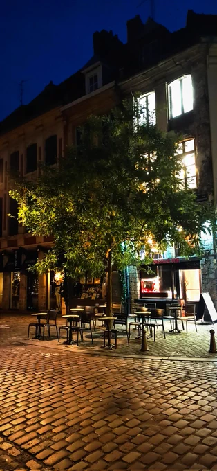 Cette photo illustre une rue pavée baignée dans l’ambiance feutrée d'un matin à Lille. Les lumières chaudes des vitrines et des fenêtres illuminent la façade d’un bâtiment ancien, ajoutant une touche de vie à la scène. Au premier plan, des tables et des chaises disposées en terrasse semblent inviter à une pause sous un arbre dont les feuillages sont également mis en valeur par les éclairages environnants. En arrière-plan, le ciel bleu nuit contraste avec les tons dorés des éclairages.

