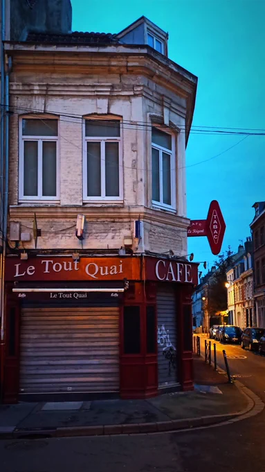 Café à l'angle de deux rues un matin à Lille. Le coin de rue est dominé par une façade du café nommé "Le Tout Quai", fermé à cette heure. Les tons clairs des murs du bâtiment, d'architecture ancienne, contrastent avec la devanture rouge et l'enseigne TABAC de l'établissement. Les lampadaires de la rue à droite éclairent les façades et les voitures garées le long du trottoir. Les tons jaunes de l'eclairage sont mis en evidence par la couleur d'un ciel bleu clair. 

