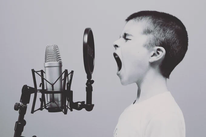 A child screaming into a microphone with a pop-filter in front of it. The picture is a black and white photography taken so that the boy and microphone is viewed from the side. 