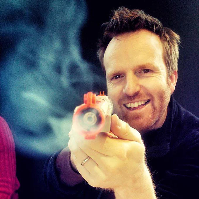 A white man with short brown hair smiles and raises a small smoking laser device at the camera. 
