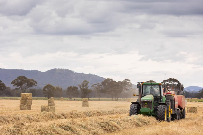 A John Deere tractor operating in a field, towing equipment and surrounded by bales of hay, with trees and overcast sky in the background.