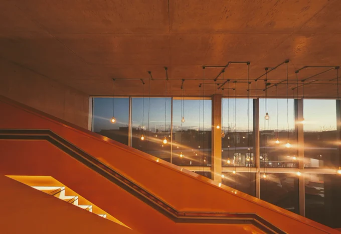 Photo taken indoors of an orange staircase and concrete ceiling against a sunset 